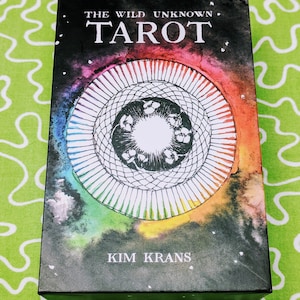 Może przedstawiać: Pudełko talii kart tarota The Wild Unknown ma czarne tło z kolorowym wzorem akwarelowym w kształcie koła z białym środkiem i tęczową obw&oacute;dką. Tekst "The Wild Unknown Tarot" jest nadrukowany na g&oacute;rze pudełka białym kolorem, a imię "Kim Krans" jest nadrukowane białym kolorem na dole.