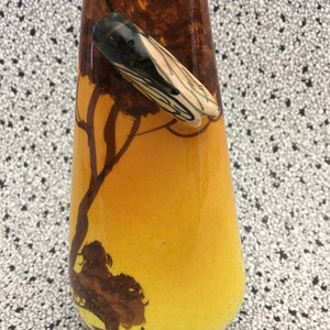 May include: A yellow and brown ceramic vase with a silhouette of a tree and a fence. A small, white and black insect is perched on the tree.