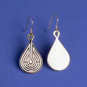 May include: A pair of gold-tone drop earrings with a white enamel maze design. The earrings have a teardrop shape and are suspended from a small hook.