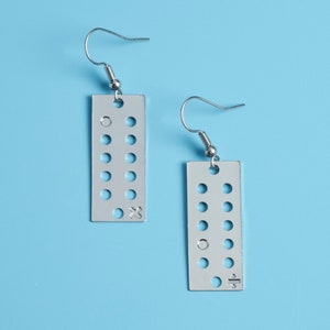 May include: A pair of silver earrings with a rectangular design. Each earring has a series of small circles and a symbol at the bottom. The symbol on the left earring is a cross, and the symbol on the right earring is a division sign.