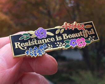 May include: A rectangular black enamel pin with gold trim and the words "Resistance is Beautiful" in white script. The pin is decorated with colorful floral accents in purple, red, and blue. The pin is held in a person's hand.