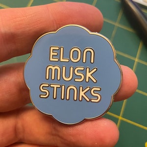 May include: A blue enamel pin in the shape of a cloud with the text "ELON MUSK STINKS" in white outlined letters.