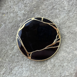 May include: A black circular pin with a gold outline and a crackled design.