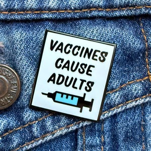May include: A square enamel pin with the text "VACCINES CAUSE ADULTS" in black, with a syringe graphic. The pin has a white background and a black border. The pin is set against a blue denim background.