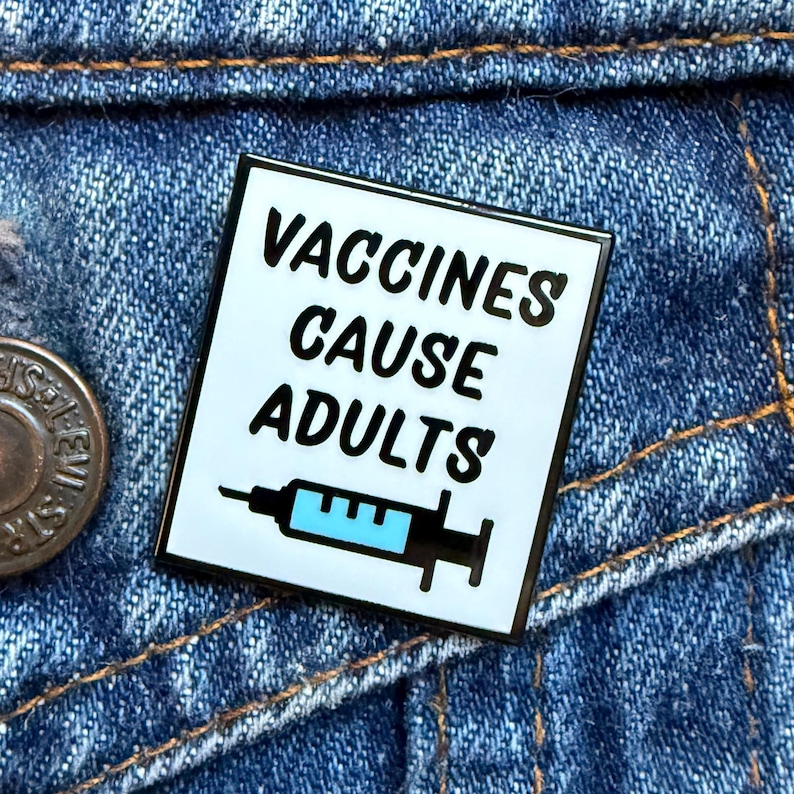 May include: A square enamel pin with the text "VACCINES CAUSE ADULTS" in black, with a syringe graphic. The pin has a white background and a black border. The pin is set against a blue denim background.