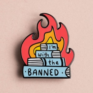 May include: Enamel pin featuring a stack of books engulfed in flames. The pin has a black outline with red, orange, and yellow flames. The books are light blue with the words "I'm with the BANNED" in white.