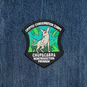 May include: Black and green embroidered patch with a white creature with red eyes. The patch reads "Cryptid Conservation Corps" and "Chupacabra Reintroduction Program".