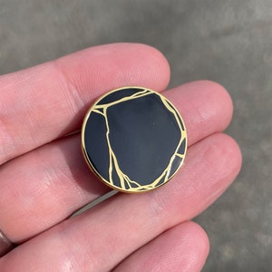 May include: A round, black enamel pin with a gold-colored border and gold-colored crack-like lines. The pin is held in a person's hand. The pin is a decorative accessory.