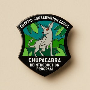 May include: Black and green enamel pin with a white creature with red eyes and a tail. The pin reads "Cryptid Conservation Corps" and "Chupacabra Reintroduction Program".