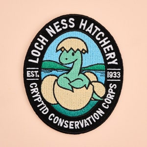 May include: A black and white embroidered patch with a green dinosaur hatching from an egg. The patch reads "Loch Ness Hatchery | Est. 1933 | Cryptid Conservation Corps".