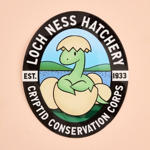May include: A round black sticker with the text "LOCH NESS HATCHERY CRYPTID CONSERVATION CORPS" in white. The sticker features a cartoon Loch Ness Monster hatching from an egg, with a blue lake and green hills in the background. The text "EST. 1933" is also included.