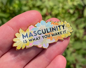 Puede incluir: Un pin de esmalte dorado con el texto "MASCULINITY IS WHAT YOU MAKE IT" en letras negras. El pin está rodeado de flores coloridas en tonos amarillos, azules, naranjas y morados. El pin se sostiene en una mano sobre un fondo verde borroso.