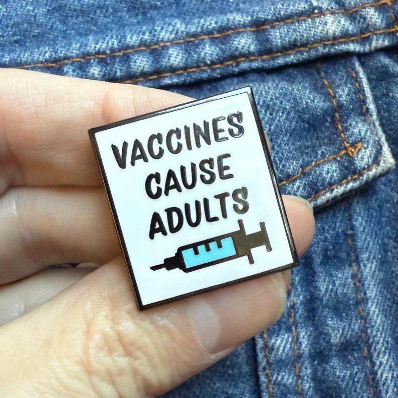 May include: A square enamel pin with a white background and black border. The pin features the text "VACCINES CAUSE ADULTS" in black, with a blue syringe graphic below. The pin is set against a denim background.