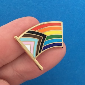 May include: A gold-colored pin in the shape of a flag with a rainbow design. The flag has seven stripes in the colors red, orange, yellow, green, blue, indigo, and violet. The flag also has a black and brown chevron pattern in the upper left corner.