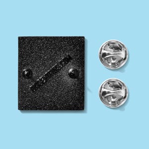 May include: A black square enamel pin with two pin backs and two silver pin clutches. The pin has a textured surface and the text "dress2impress.com" printed diagonally across the back.