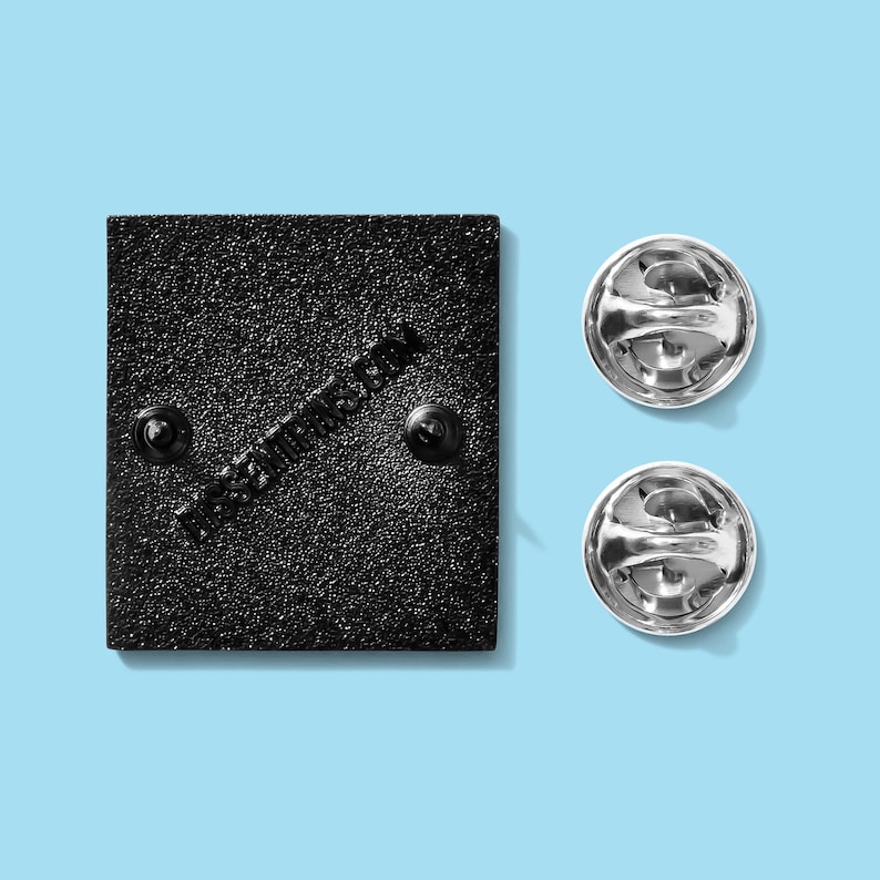 May include: A black square enamel pin with two pin backs and two silver pin clutches. The pin has a textured surface and the text "dress2impress.com" printed diagonally across the back.