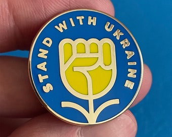 May include: A blue and yellow enamel pin with the text "Stand With Ukraine" and a stylized yellow fist in the center.