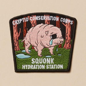 May include: Embroidered patch featuring a pink, crying Squonk creature from the Cryptid Conservation Corps. The patch has a black border, green grass, and red-brown trees. Text includes "CRYPTID CONSERVATION CORPS" and "SQUONK HYDRATION STATION."