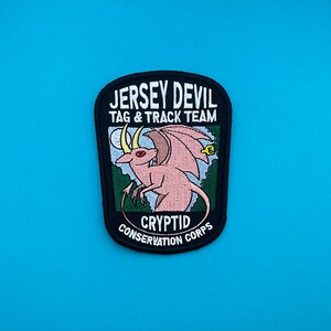 May include: Black and white embroidered patch with the text "Jersey Devil Tag & Track Team" and "Cryptid Conservation Corps". The patch features a pink, winged creature with red eyes and a yellow tag.