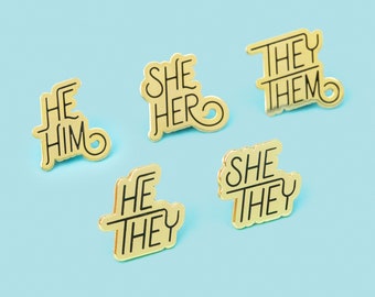 May include: Six gold-colored pins with black lettering. The pins feature different pronoun combinations, including "He Him", "She Her", "They Them", "He They", and "She They".