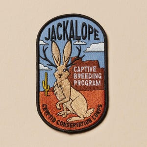 May include: Embroidered patch featuring a jackalope with antlers, set against a desert landscape with a blue sky. The patch includes the text "JACKALOPE", "CAPTIVE BREEDING PROGRAM", and "CRYPTID CONSERVATION CORPS".