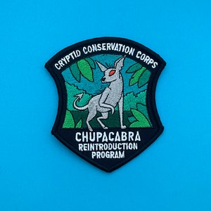 May include: Black and white embroidered patch with a white creature with red eyes standing in a green jungle. The patch reads "Cryptid Conservation Corps" and "Chupacabra Reintroduction Program".