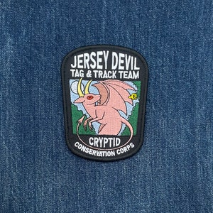 May include: A patch featuring the text "Jersey Devil Tag & Track Team Cryptid Conservation Corps." The patch has a black border and depicts a pink Jersey Devil creature with yellow horns and wings. The background is blue.