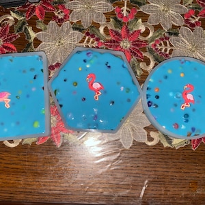 May include: Three blue resin coasters with a white border and pink flamingo accents. The coasters are shaped like a square, hexagon, and circle.