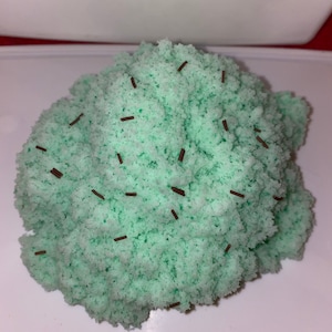 May include: A mound of light mint green textured sensory dough or slime with a porous, crumbly appearance. The surface is scattered with small, thin, dark brown cylindrical sprinkles that are distributed unevenly across the mint green mass.
