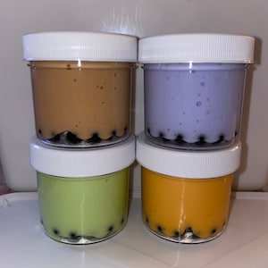 May include: Four clear plastic jars with white lids, each filled with a different colored slime. The colors are brown, purple, green, and orange. Black beads are visible at the bottom of each jar, resembling boba.