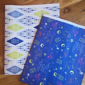 May include: Two sheets of wrapping paper with different designs. One sheet is white with a blue and yellow geometric pattern and the text "Ramadan Mubarak". The other sheet is blue with a white pattern of stars, camels, palm trees, and crescent moons. The text "Ramadan Kareem" is repeated throughout the pattern.