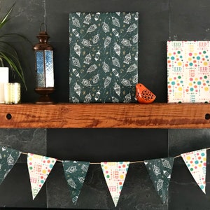 Puede incluir: Un papel estampado verde y dorado con linternas blancas y estrellas. Un papel blanco y colorido con la palabra "Eid" repetida. Una guirnalda de banderas triangulares con la palabra "Eid" y patrones coloridos. Una peque&ntilde;a figurita de p&aacute;jaro naranja.