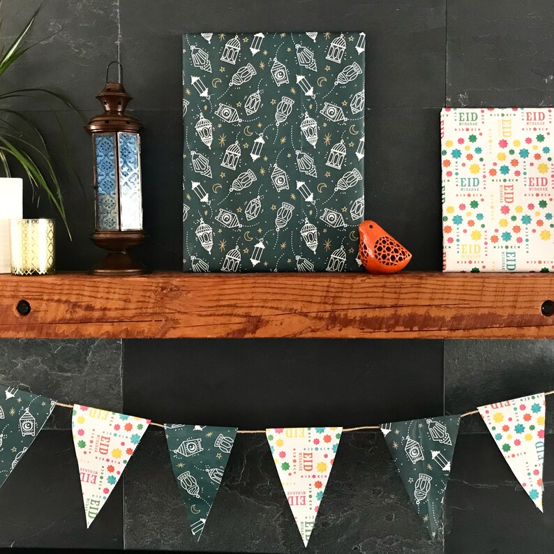Puede incluir: Un papel estampado verde y dorado con linternas blancas y estrellas. Un papel blanco y colorido con la palabra "Eid" repetida. Una guirnalda de banderas triangulares con la palabra "Eid" y patrones coloridos. Una peque&ntilde;a figurita de p&aacute;jaro naranja.