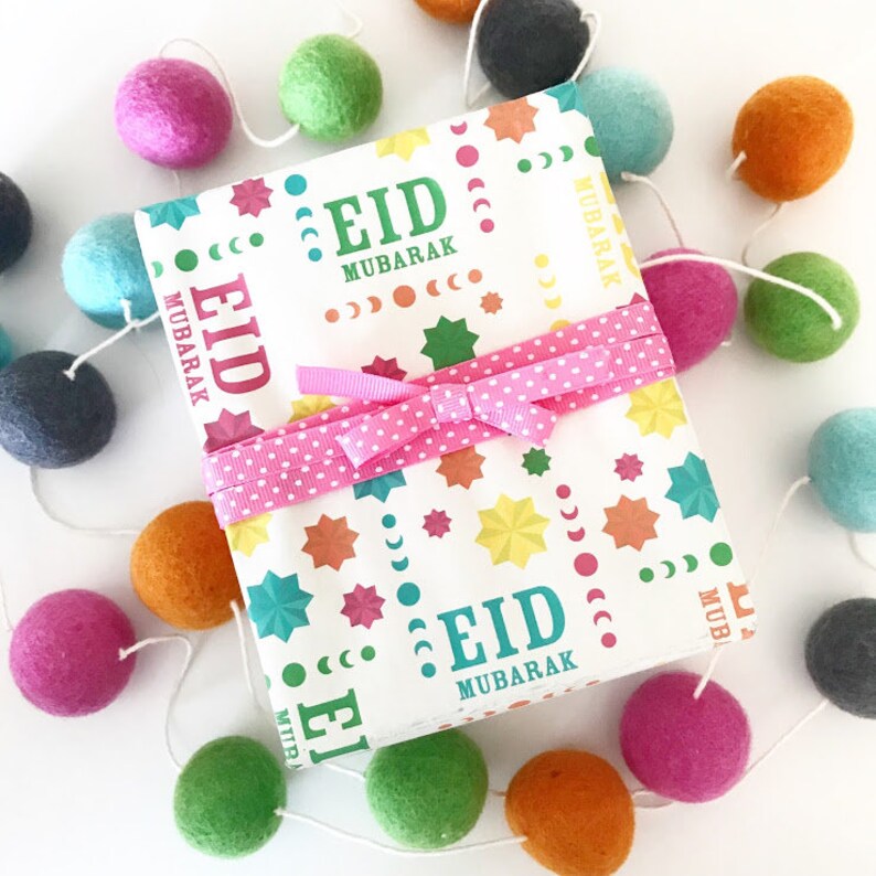 Puede incluir: Una caja de regalo blanca con una cinta rosa y un patr&oacute;n colorido de estrellas y medias lunas. La caja est&aacute; envuelta en papel que dice "Eid Mubarak" en una fuente azul verdosa. La caja est&aacute; rodeada de una guirnalda de bolas de fieltro de colores.