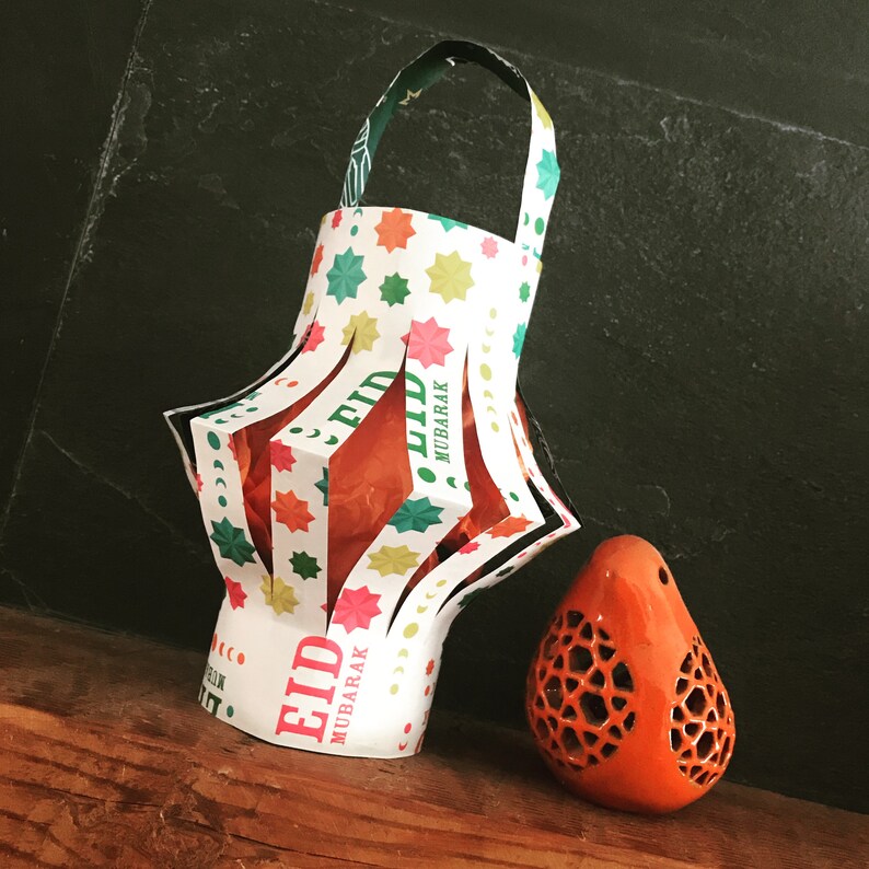 Puede incluir: Una linterna de papel decorada con estrellas de colores y las palabras "Eid Mubarak". La linterna est&aacute; hecha de papel blanco con detalles verdes, rosas y naranjas. Una peque&ntilde;a linterna de cer&aacute;mica naranja se encuentra delante de la linterna de papel.