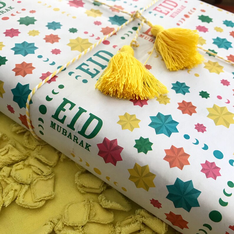 Puede incluir: Papel de regalo blanco con un patr&oacute;n de estrellas de colores y el texto "Eid Mubarak". El papel est&aacute; atado con una cuerda blanca y amarilla y decorado con dos borlas amarillas.