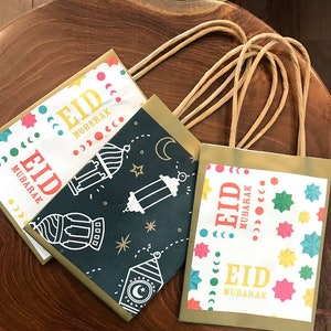 Puede incluir: Tres bolsas de regalo doradas con dise&ntilde;os coloridos y el texto "Eid Mubarak" en cada bolsa. Las bolsas tienen asas de papel marr&oacute;n.