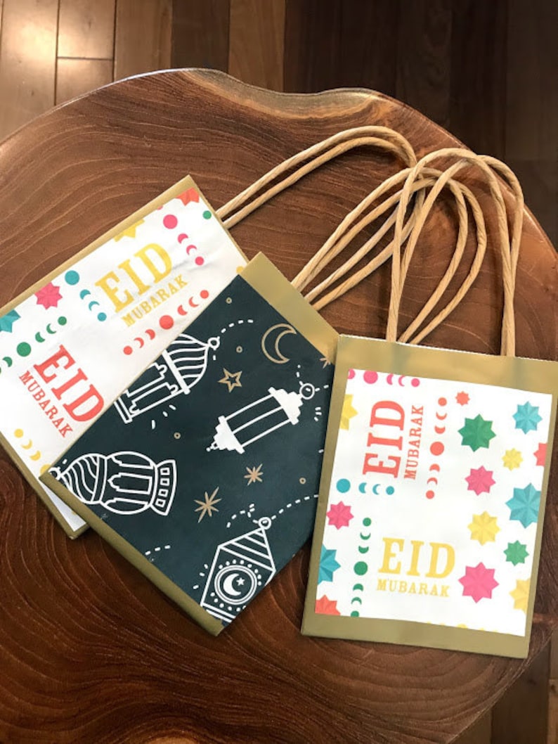 Puede incluir: Tres bolsas de regalo doradas con dise&ntilde;os coloridos y el texto "Eid Mubarak" en cada bolsa. Las bolsas tienen asas de papel marr&oacute;n.