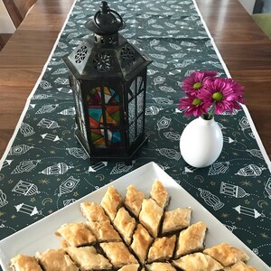 Puede incluir: Una mesa puesta con un mantel verde con un patr&oacute;n de linternas. Una linterna de metal negro con paneles de vidrio coloreado se encuentra sobre la mesa. Un jarr&oacute;n blanco con flores rosas se encuentra sobre la mesa. Un plato blanco con una variedad de baklava se encuentra sobre la mesa.