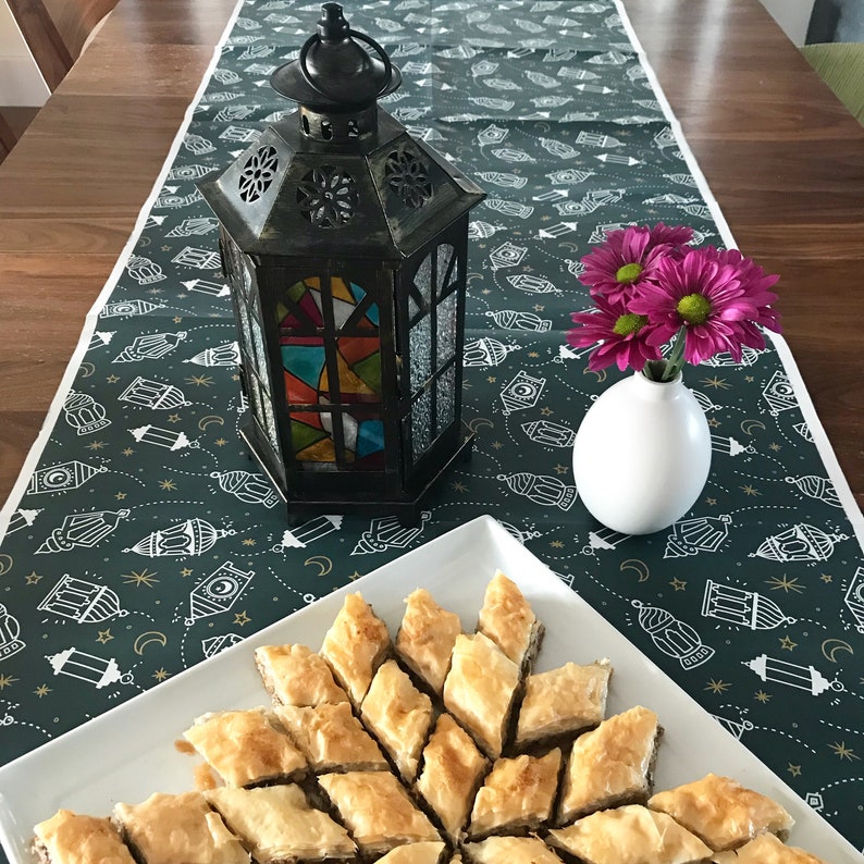 Puede incluir: Una mesa puesta con un mantel verde con un patr&oacute;n de linternas. Una linterna de metal negro con paneles de vidrio coloreado se encuentra sobre la mesa. Un jarr&oacute;n blanco con flores rosas se encuentra sobre la mesa. Un plato blanco con una variedad de baklava se encuentra sobre la mesa.