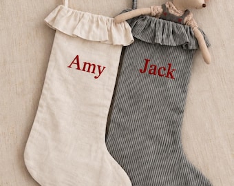 May include: Two fabric Christmas stockings with ruffled tops and hanging loops. One stocking is solid cream with the name Amy embroidered in red. The other is grey pinstriped with the name Jack in red, featuring a plush mouse toy wearing a red hat.