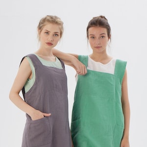 May include: Two women wearing linen aprons. The apron on the left is a dark gray color and the apron on the right is a light green color. Both aprons have a single pocket on each side.