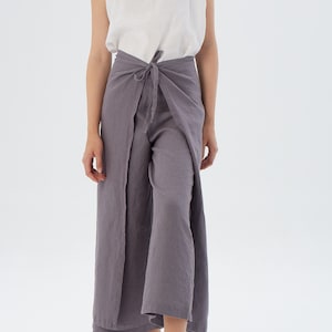 Puede incluir: Una mujer con una camiseta sin mangas blanca y pantalones envolventes grises. Los pantalones se atan a la cintura y tienen un dise&ntilde;o de pernera ancha. El conjunto est&aacute; hecho de una tela ligera, perfecto para el clima c&aacute;lido. El fondo es blanco liso.