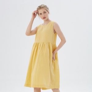 May include: A sleeveless, yellow linen dress with a gathered waist and pockets. The dress falls to the mid-calf and has a simple, round neckline. The model is wearing the dress in a studio setting with a white background.