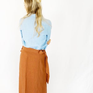 May include: A rust-colored linen wrap skirt with a long, flowing silhouette. The skirt is tied at the waist with a fabric sash.