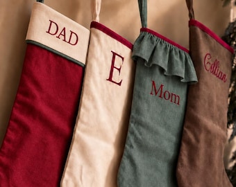 May include: Four hanging Christmas stockings in red, cream, sage green, and brown fabric. The red stocking has a cream cuff labeled DAD. The cream stocking features the letter E. The sage green stocking has a ruffled top and is labeled Mom. The brown stocking is labeled Gillian.