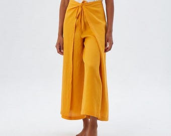 May include: Mustard yellow wide-leg trousers featuring a wrap-style front panel with a tie-waist closure. The garment has a textured fabric finish and falls to a cropped length, paired with a simple white sleeveless top.