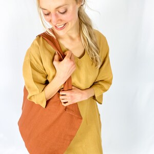 May include: A woman wearing a mustard yellow linen dress holds a large, rust-colored tote bag.