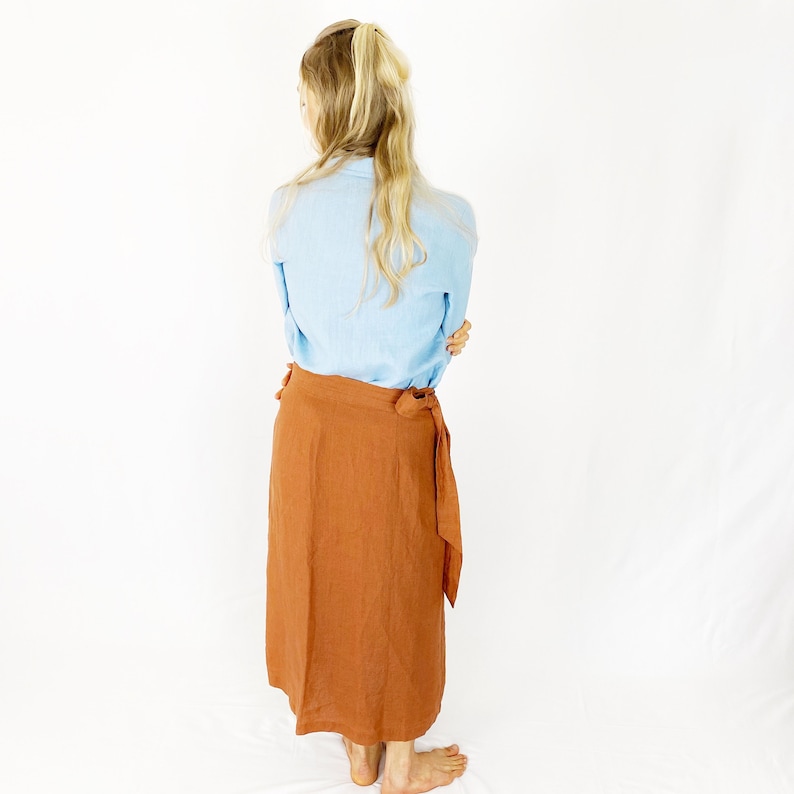 May include: A rust-colored linen wrap skirt with a long, flowing silhouette. The skirt is tied at the waist with a fabric sash.