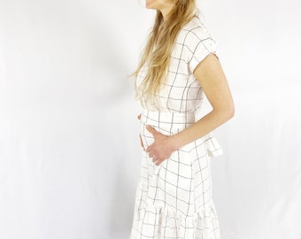 May include: A white midi dress featuring a black grid pattern, short sleeves, a fitted waistline, and a tiered skirt with a ruffled hem. The garment is constructed with a simple bodice and a tie detail visible at the back waist area.